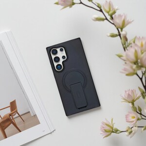 May include: A black phone case with a built-in stand and a circular magnetic attachment. The case has a camera cutout and is lying on a white surface next to a book and pink flowers.