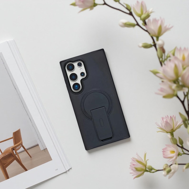 May include: A black phone case with a built-in stand and a circular magnetic attachment. The case has a camera cutout and is lying on a white surface next to a book and pink flowers.