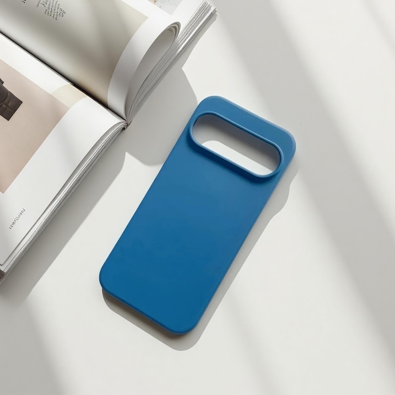 May include: A solid blue phone case with a rectangular design and a large camera cutout. The case is lying on a white surface next to an open magazine.