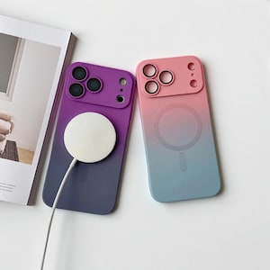 Puede incluir: Dos fundas de teléfono con degradado. Una funda pasa de morado a azul oscuro, la otra de rosa a azul claro. Ambas fundas tienen una base de carga blanca circular y un módulo de cámara.