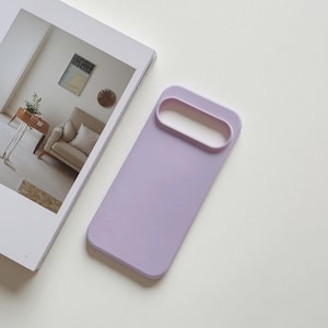 May include: A lavender phone case with a rectangular design and a large, oval camera cutout. The case is lying on a white surface next to an open magazine with a picture of a living room.