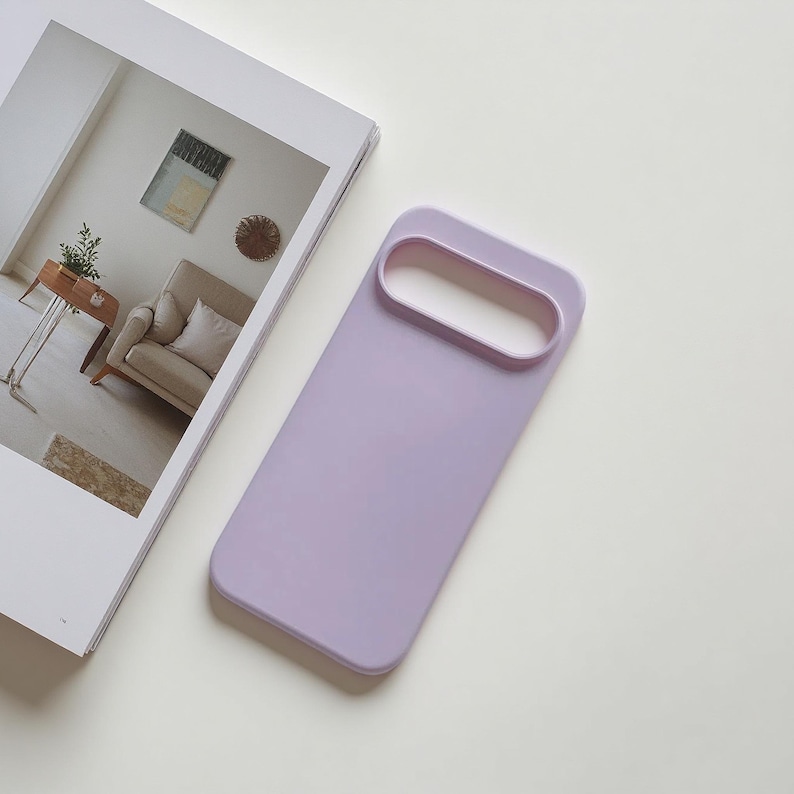 May include: A lavender phone case with a rectangular design and a large, oval camera cutout. The case is lying on a white surface next to an open magazine with a picture of a living room.