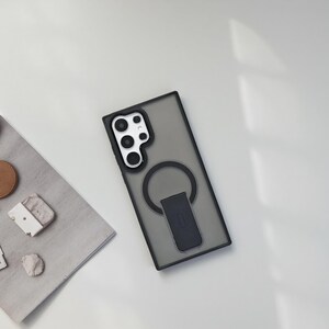 May include: A black and gray phone case with a ring holder. The case has a clear, smoky appearance and a black border. The ring holder is black and has a rectangular base. The phone case is on a white surface.