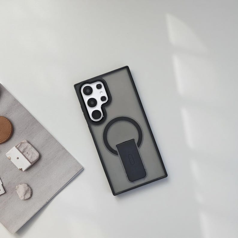 May include: A black and gray phone case with a ring holder. The case has a clear, smoky appearance and a black border. The ring holder is black and has a rectangular base. The phone case is on a white surface.