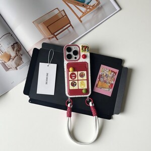 May include: A white and red phone case with a candy-themed design, featuring colorful accents and a matching lanyard. The case has a playful aesthetic with a unique, eye-catching appearance. The tag reads "INS HOUSE".