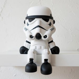 May include: A white and black Stormtrooper figurine with a rounded head, black accents, and black feet. The figurine is sitting on a white shelf against a textured white wall. The design is inspired by the Star Wars franchise.