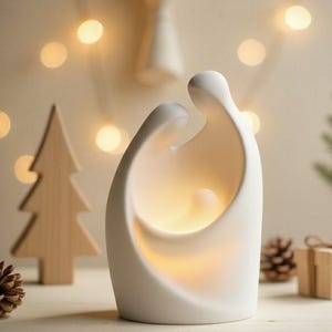 May include: A white ceramic nativity scene with a soft, glowing light emanating from within. The minimalist design features two figures embracing, with a third figure nestled between them. A wooden Christmas tree and pinecone are in the background.