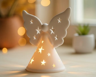 May include: A white ceramic angel figurine with star-shaped cutouts on the wings and cone-shaped body. The angel is illuminated from within, creating a warm glow. The wings are detailed with multiple star cutouts. The figurine is a decorative item.