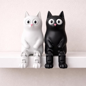 Op de afbeelding: Een paar kattenbeeldjes, één wit en één zwart, zitten naast elkaar op een witte plank. Elke kat heeft grote, ronde ogen, een roze neus en zit met zijn benen te bungelen. De achtergrond is een lichtroze muur.