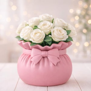 May include: A bouquet of white roses with green leaves in a pink, round vase. The vase has a bow detail and a ruffled top. The background is softly blurred with bokeh lights.
