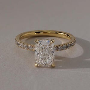 May include: A gold engagement ring with a large, rectangular-cut diamond as the centerpiece. The band is also adorned with smaller diamonds, creating a sparkling effect. The ring is set against a neutral background.