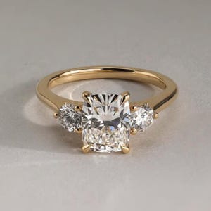 Può includere: Un anello d'oro con un grande diamante centrale a taglio rettangolare, affiancato da due diamanti rotondi più piccoli. L'anello ha una fascia semplice, che mette in risalto la brillantezza delle pietre preziose. L'anello è su uno sfondo neutro.