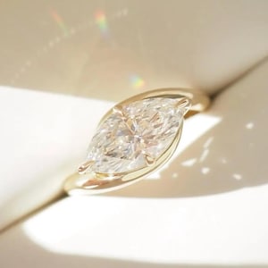 Peut inclure: Une bague en or ornée d'un diamant taille marquise. Le diamant est serti dans une monture à lunette, et le corps de la bague est en or poli. La bague est présentée dans un écrin blanc, avec un éclairage doux mettant en valeur les facettes du diamant.