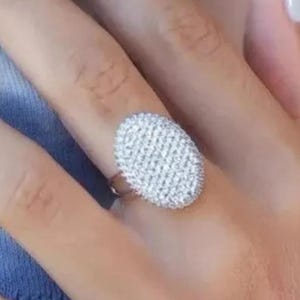 Könnte beinhalten: Ein silberner Ring mit einer ovalen, mit kleinen, funkelnden Steinen besetzten Oberfläche. Der Ring befindet sich an einer Hand mit hellblauem Nagellack. Der Hintergrund ist ein hellblau-weißes Batikmuster.