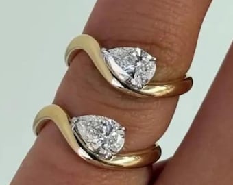 May include: Two gold rings with a pear-shaped diamond set in a wave design. The rings are on a finger, showcasing the jewelry's sparkle and elegant style. The gold band curves around the diamond, creating a modern look.