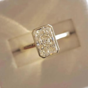 Puede incluir: Un anillo de oro con una piedra preciosa rectangular y transparente engastada en un bisel. El anillo se presenta en una caja blanca, con una banda delgada. La piedra preciosa es el punto focal, captando la luz y mostrando su claridad.