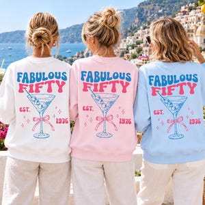 Op de afbeelding: Drie sweaters met ronde hals in wit, lichtroze en lichtblauw, elk met een achteropdruk van een martiniglas met olijven en een strik. De tekst luidt FABULOUS FIFTY EST. 1976 in blauwe en roze retro-stijl letters met kleine sterretjes als accent.