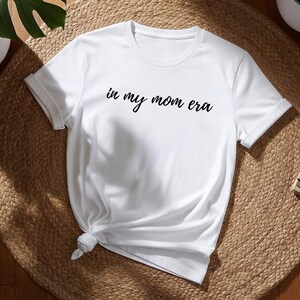 Könnte beinhalten: Weißes T-Shirt mit dem Schriftzug "in my mom era" in schwarzer Schrift. Das Shirt ist unten geknotet und liegt auf einer runden, gewebten Matte. Topfpflanzen und Bücher sind im Hintergrund.