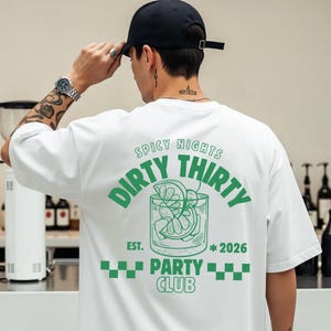 Op de afbeelding: Wit T-shirt met een groene afbeelding op de rug met de tekst "Dirty Thirty Party Club" en een cocktailillustratie. Het shirt heeft korte mouwen en een ronde hals. De persoon draagt een zwarte baseball cap.