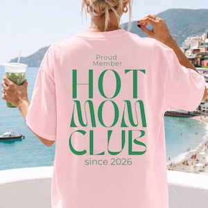 Op de afbeelding: Lichtroze T-shirt met de groene tekst "Proud Member HOT MOM CLUB since 2026". De persoon die het shirt draagt heeft blond haar, met een uitzicht op zee op de achtergrond.