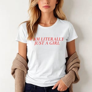 Könnte beinhalten: Weißes T-Shirt mit dem roten Text "I AM LITERALLY JUST A GIRL". Das Shirt hat einen Rundhalsausschnitt und kurze Ärmel. Ein beigefarbener Cardigan ist über den Schultern drapiert.