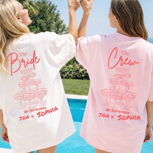 Könnte beinhalten: Zwei übergroße T-Shirts, eines weiß und eines rosa, mit passendem rotem Text und Grafiken. Das weiße Shirt trägt die Aufschrift "The Bride" und das rosa Shirt "The Crew". Beide Shirts zeigen eine Champagnerturm-Grafik und ein Hochzeitsdatum.