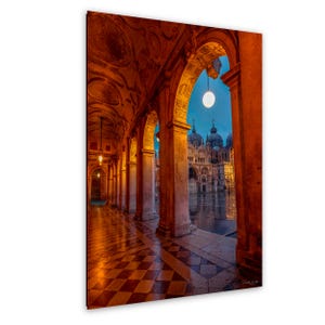 May include: A photograph of a Venetian colonnade at dusk. The image features a series of arches and columns, with a checkered floor leading towards a view of a cathedral. The sky is a deep blue, and the scene is illuminated by warm, golden light.