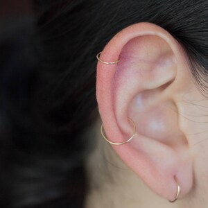 Pu&ograve; includere: Primo piano di un orecchio con tre orecchini a cerchio d'oro. Gli orecchini sono nei piercing dell'elice, della conchiglia e del lobo.