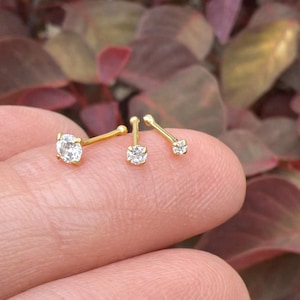 May include: Three gold-tone nose studs, each featuring a single clear round-cut gemstone set in a prong setting. The studs vary in size, with the largest on the left and the smallest on the right, all attached to thin, straight gold-tone metal posts.