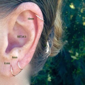 Pu&ograve; includere: Set di tre orecchini a cerchio in argento di diverse dimensioni: 6 mm, 8 mm e 10 mm. Gli orecchini sono mostrati nell'orecchio di una persona.