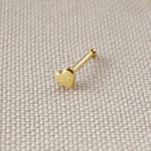 Könnte beinhalten: Ein goldfarbener Nasenstecker mit einem kleinen, polierten herzförmigen Kopf, der an einem geraden, schlanken Stift befestigt ist. Das Schmuckstück hat eine glatte, reflektierende Metalloberfläche und ist für Piercings konzipiert.