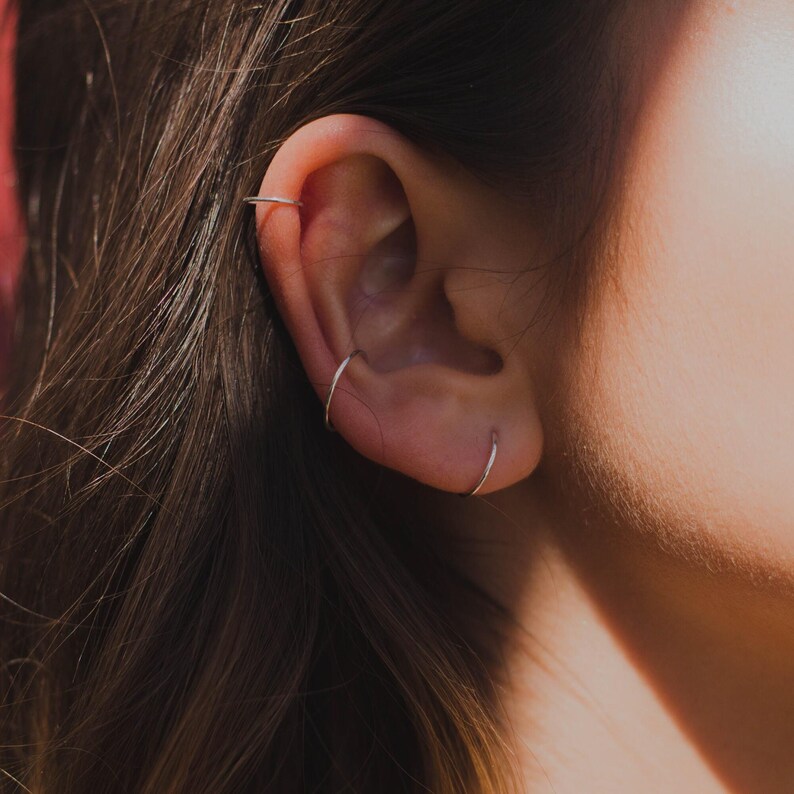 Pu&ograve; includere: Una persona indossa tre piccoli anelli d'argento all'orecchio. Gli orecchini sono nei piercing dell'elice, del conch e del lobo.
