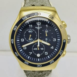 Op de afbeelding: Goudkleurig Swatch chronograaf horloge met een zwarte wijzerplaat en witte markeringen. Het horloge heeft drie sub-wijzerplaten, een datumvenster en een gestructureerde grijze band. De bezel heeft zwarte markeringen voor minuten en seconden.