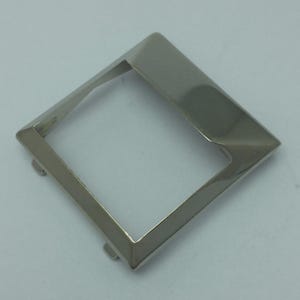 Puede incluir: Un marco cuadrado de metal plateado brillante con un borde biselado. El marco tiene forma de diamante con un centro hueco y pequeñas pestañas en dos esquinas. El objeto es probablemente un componente decorativo o funcional.