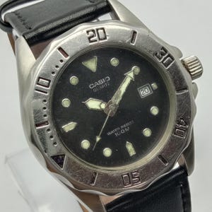 May include: A Casio Quartz wristwatch with a black dial, luminous hands, and hour markers. The watch features a silver-tone rotating bezel with engraved numbers, a date window at the three o'clock position, and a black leather strap.