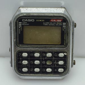May include: A vintage Casio CA-86 calculator watch with a silver-tone case. The watch features a calculator keypad, a digital display, and the Casio and CA-86 model names. The watch has buttons for mode, light, adjust, and start/stop functions.