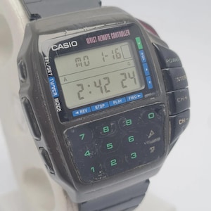 Op de afbeelding: Een vintage Casio Wrist Remote Controller horloge. Het horloge heeft een zwarte rechthoekige wijzerplaat met een digitale display die de tijd en datum weergeeft. Het heeft knoppen voor verschillende functies, waaronder volume, kanaalbediening en afstandsbediening.