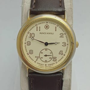 May include: A gold-toned wristwatch with a cream-colored face and brown leather band. The watch face features black numerals, hands, and the text "PRINCE HOTELS". A smaller dial is at the bottom. The watch is a classic style.
