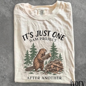 May include: A cream-colored short-sleeve t-shirt featuring a brown beaver holding a log next to a woodpile and pine trees. The text reads IT'S JUST ONE DAM PROJECT AFTER ANOTHER. The shirt has a Comfort Colors brand label at the inner neckline.