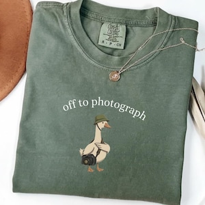 Könnte beinhalten: Ein olivgrünes T-Shirt mit Rundhalsausschnitt und einer Grafik einer Gans, die einen Hut trägt und eine Kamera bei sich hat, sowie dem in Weiß gedruckten Text "off to photograph". Das T-Shirt hat ein Comfort Colors Markenetikett am Kragen.