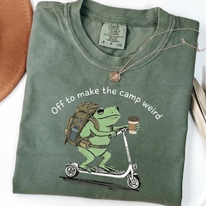 Könnte beinhalten: Grünes T-Shirt mit einem Frosch-Motiv, der Roller fährt, einen Rucksack und einen Kaffeebecher trägt. Der Text lautet: "Off to make the camp weird." Das Shirt hat ein Comfort Colors-Etikett und eine silberne Halskette.