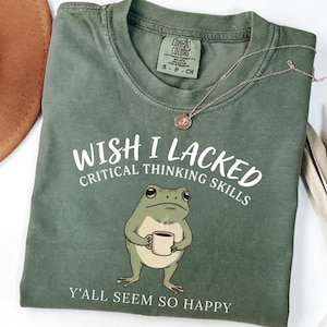 Könnte beinhalten: Ein olivgrünes T-Shirt mit einer Grafik eines Frosches, der eine Kaffeetasse hält. Der Text lautet WISH I LACKED CRITICAL THINKING SKILLS und Y'ALL SEEM SO HAPPY. Das T-Shirt hat einen Rundhalsausschnitt und ein Comfort Colors Etikett am Innenkragen.
