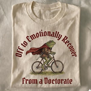 Puede incluir: Camiseta color crema con una ilustración caprichosa de una rana montando en bicicleta, con capa y espada. El texto "Off to Emotionally Recover From a Doctorate" está impreso en borgoña, arqueado sobre y debajo de la imagen.