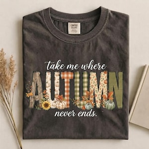 May include: A charcoal gray crew neck t-shirt featuring the text "take me where AUTUMN never ends." The word "AUTUMN" is displayed in large block letters filled with various patterns including plaid, sunflowers, and pumpkins in orange and green.