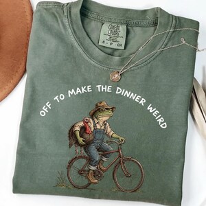 Puede incluir: Una camiseta de color verde oliva con un gráfico de una rana con sombrero y peto montada en bicicleta y sosteniendo un pavo. El texto sobre el gráfico dice: "OFF TO MAKE THE DINNER WEIRD." La camiseta tiene una etiqueta de la marca Comfort Colors.