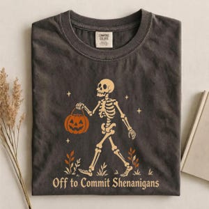 May include: A charcoal grey crew neck t-shirt featuring a graphic of a beige skeleton walking while carrying an orange jack-o'-lantern. The design includes small stars and foliage, with the text "Off to Commit Shenanigans" printed below.