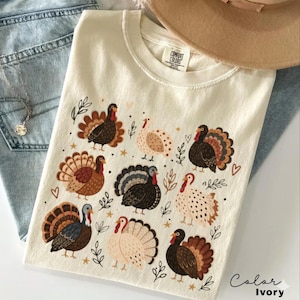 Puede incluir: Una camiseta color marfil con etiqueta Comfort Colors que presenta un estampado gráfico repetitivo de varios pavos en tonos marrón, beige y negro, intercalados con pequeñas ramas de hojas, estrellas y corazones en la parte delantera.