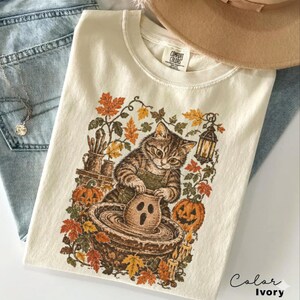 以下が含まれることがあります： アイボリー色のTシャツ。ろくろを使ってお化けの形の粘土ポットを作るキジトラ猫のグラフィックプリントが施されており、周囲には秋の葉、2つのジャック・オー・ランタン、吊り下げ式のランタン、火のついたキャンドルが描かれています。