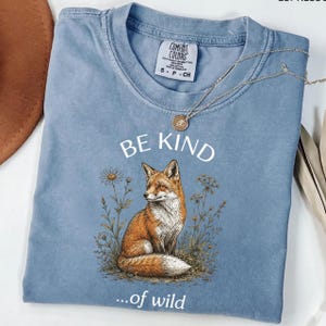 Peut inclure: Un t-shirt bleu poussiéreux à manches courtes avec un motif représentant un renard roux assis parmi des fleurs sauvages. Le texte "BE KIND" est imprimé en blanc au-dessus du renard, et "...of wild" est imprimé en blanc en dessous. Un collier doré.