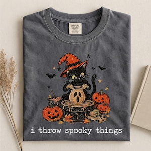 May include: A charcoal gray t-shirt featuring a graphic of a black cat wearing an orange witch hat, working at a pottery wheel with a ghost-faced clay pot. The design includes two jack-o'-lanterns, bats, stars, and the text i throw spooky things.